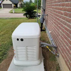 A beige Generac generator is shown on a concrete pad, next to a red brick house. It is connected to the house's utility system with several pipes and wires. The generator has a sloped top and side vents.