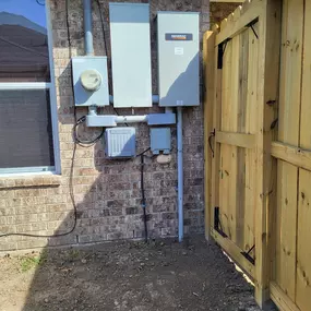 An outdoor electrical setup on the side of a house with a red brick wall. The installation includes a Generac generator, an electric meter, and other grey utility boxes and conduit pipes.