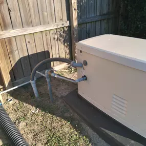 A side view of a Generac generator installed on a concrete slab against a wooden fence. Gray pipes and electrical conduits are connected to the unit, with one pipe leading into the ground.