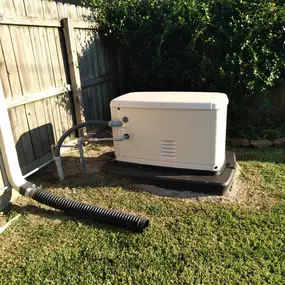 An image of a Generac home backup generator on a concrete pad, situated in a grassy yard. The generator is next to a wooden fence and a large, green hedge. Pipes and a black corrugated drain pipe are also visible.