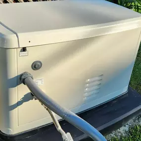 A close-up shot of a Generac home generator, showcasing its beige casing, vents, and a gray pipe connected to its side. The unit rests on a black, raised foundation on a patch of grass.