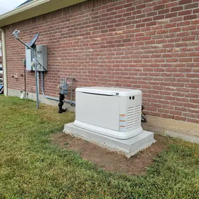 A beige Generac generator is installed on a concrete pad in a residential yard. The unit is placed against a red brick wall of a house, with an electrical meter and other utility equipment nearby.