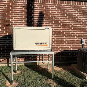 A beige Generac generator is mounted on a metal stand against a red brick wall. An air conditioning unit is visible to the right of the generator, with both installations casting shadows on the wall.