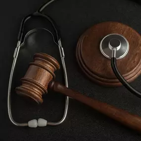 A wooden gavel and a stethoscope are arranged on a dark, textured surface, representing the intersection of law and medicine.