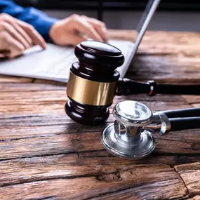 A gavel and stethoscope are placed on a rustic wooden desk, with a person's hands typing on a laptop in the blurred background.
