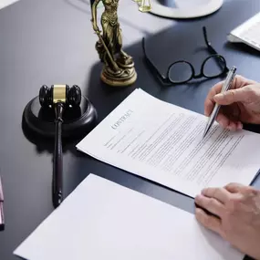 A lawyer in a blue shirt is signing a contract at a black desk, with a gavel, statue of Lady Justice, and a law book visible.