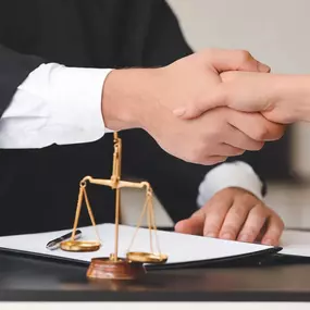 Two individuals shake hands over a dark desk with the golden scales of justice prominently displayed, suggesting a legal agreement or partnership.