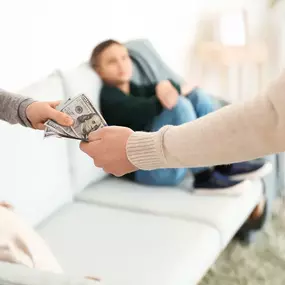 A close-up of two people's hands exchanging money, with a child sitting on a couch in the blurry background, representing child support or custody payments.