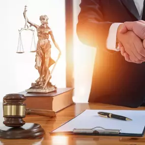 Two individuals in formal attire shake hands over a wooden table, with a gavel and a statue of Lady Justice in the foreground, signifying a legal settlement.