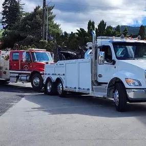 A large white tow truck is hitched to a red fire truck, parked on asphalt with trees in the background.
