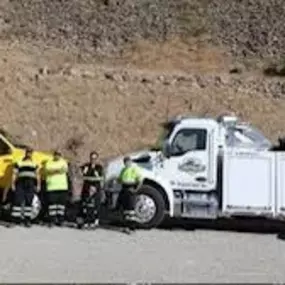A line of four tow trucks—white van, yellow, white, and black—are parked side-by-side with two crew members.