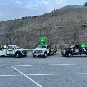 Three tow trucks, one decorated with a large green leprechaun hat, are parked in a lot against a mountain backdrop.