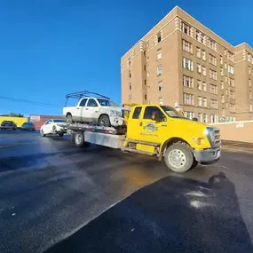 A yellow flatbed tow truck carries a white pickup truck, with a smaller white car towed behind, on a sunny day.