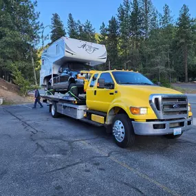 A yellow flatbed tow truck carries a dark pickup truck with a large white camper shell, parked in a lot.
