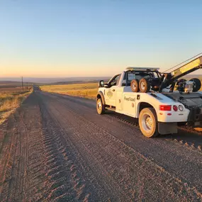 A white tow truck is parked on a dirt road, facing away, with a vast, flat landscape and clear sky in the distance.