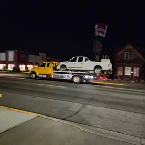 A yellow flatbed tow truck carries a white pickup truck on a street at night, with buildings and a KFC sign visible.