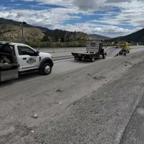 A white tow truck and a flatbed truck are on the side of a highway, with mountains in the background under a cloudy sky.