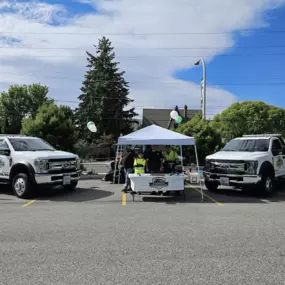 A white tent is set up between two white tow trucks in a parking lot, with people and trees around.