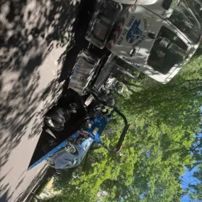 A tow truck is lifting a blue car onto its flatbed on a tree-lined street under clear, sunny weather.