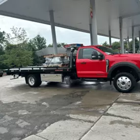 A red flatbed tow truck is parked at a gas station, with trees, buildings, and wet pavement visible around it.