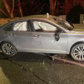 A silver sedan is badly damaged with its airbags deployed. The car sits on a tow dolly at night, likely after an accident, as two people stand nearby.