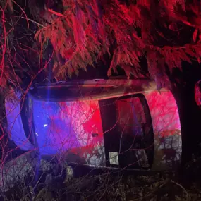 An overturned SUV is illuminated by red and blue emergency lights at night, having crashed into a dense patch of trees and bushes.