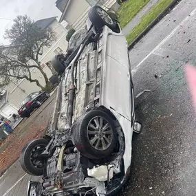 A white SUV lies upside down on a wet residential street after flipping over. The vehicle's undercarriage is exposed, with debris scattered nearby.