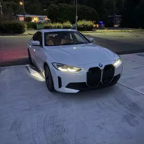 A white BMW sedan with its headlights on is parked at night in a paved lot. The car's front grille is clearly visible.