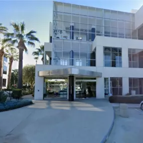 HRXCPAS Offices, a modern three-story office building with large glass windows and white exterior, located at Spectrum Business Center. Palm trees, landscaped greenery, and an American flag are visible in front, with clear blue sky and sunlight overhead.