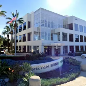Front entrance of Spectrum Business Center building, home of HRXCPAS with glass doors and white exterior, surrounded by palm trees, manicured plants, and a small parking area. The building has a sleek, contemporary design with reflective windows.