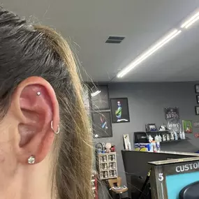 Close-up of a woman's ear showing three piercings: a stud on the lobe, a hoop in the helix, and a small stud in the upper cartilage, taken inside a tattoo studio.