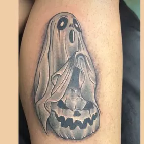 A black and gray tattoo of a ghostly figure draped in a sheet, hovering above a sinister jack-o'-lantern. The design combines eerie and Halloween-themed elements.