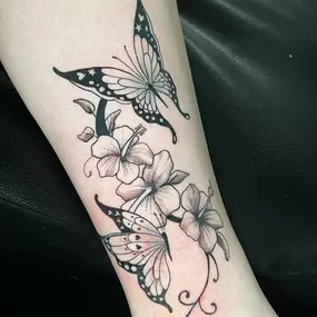 A delicate black and gray tattoo featuring two butterflies fluttering around blooming flowers. The design combines fine lines, soft shading, and ornamental details for a graceful, feminine look.