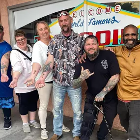 A group of six people happily show off their fresh tattoos in front of a colorful 