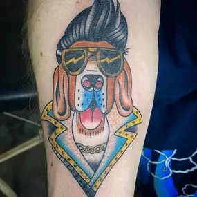 A colorful tattoo of a cartoon dog styled as Elvis Presley, complete with pompadour hair, gold sunglasses, a chain, and a flashy jacket with stars and lightning bolts.