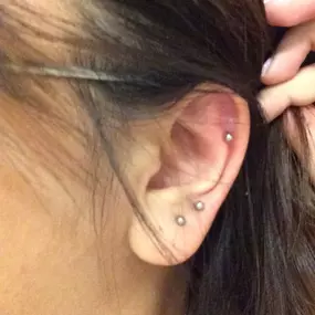 Close-up of a person's ear with three visible piercings—two on the lobe and one on the upper cartilage. The person is holding their hair back to reveal the jewelry.