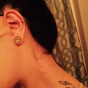 A close-up of a woman's ear featuring a helix piercing and a large decorative stud earring. Her hair is tied back, revealing a shoulder tattoo and glasses.