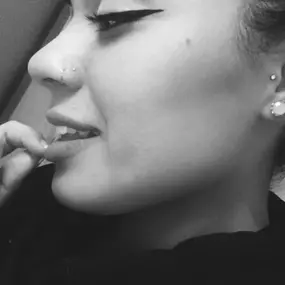 A black and white photo of a woman with multiple ear piercings, a nose stud, winged eyeliner, and manicured nails, lightly biting her finger while smiling.