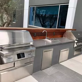 A sleek, modern outdoor kitchen area featuring stainless steel appliances. There are a large grill and a separate cooktop with a sink, all set into a gray concrete countertop with a wooden backsplash.
