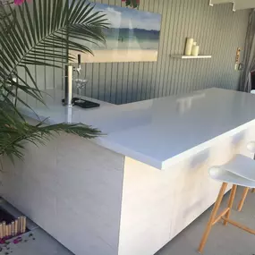 A clean, modern bar area features a white countertop and textured paneling. A beer tap and two minimalist stools are visible, with lush green plants and a framed beach photo completing the outdoor setting.