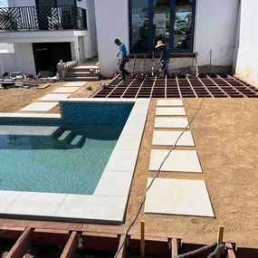 An outdoor construction site shows the early stages of a backyard transformation. Two workers are building a wooden deck frame next to a newly installed swimming pool with modern, square stepping stones leading to it.