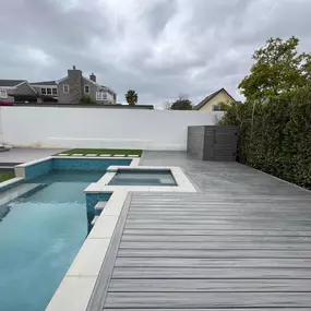A finished backyard features a gray composite deck surrounding a swimming pool with an attached spa. The modern design includes a clean white privacy wall and lush green hedges, with a cloudy sky overhead.
