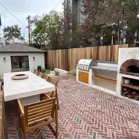 A beautifully designed outdoor patio features a herringbone-patterned brick floor. The space is set up for dining and cooking with a long concrete table, a built-in grill, and a rustic brick pizza oven.