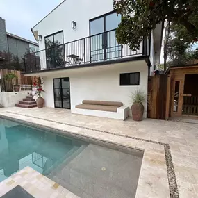 A home's outdoor area with a modern, rectangular swimming pool surrounded by beige tile paving. The space includes a built-in outdoor couch and a wooden sauna cabin, creating a resort-like atmosphere.