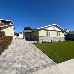 A newly renovated house with a modern, block-paved driveway leading to the garage. The front yard features a tidy, artificial grass lawn and a stone veneer on the lower half of the house's exterior.