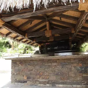 A tropical-themed outdoor bar with a thatched roof supported by textured, wood-like columns wrapped in rope. The counter features a stone base and is equipped with a modern stainless steel grill.