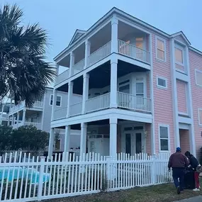 A residential HVAC unit is installed on a gray, metal-sided building. The large white door has three windows, with a table set up nearby.