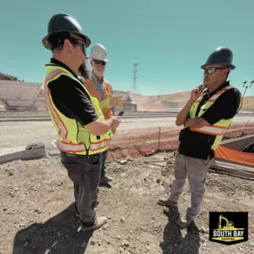 Shawn Dean South Bay Grading Three South Bay Grading (SBG) team members hold a field huddle at an active road/earthwork project. They wear hard hats, hi‑vis vests, and safety gear while discussing plans beside orange safety fencing and heavy equipment in the background. Dusty, sunny conditions with hills and power lines typical of South Bay, California. SBG logo in the bottom‑right corner.
