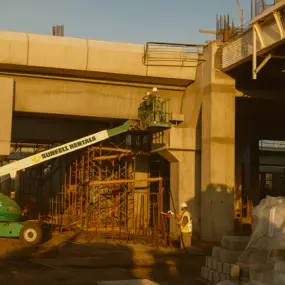 Workers performing sandblasting and painting on concrete bridge structure using boom lift and scaffolding.