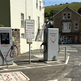 A wide view of Glen Lyn Car Park showing two InstaVolt electric vehicle chargers with visible signage and retail shops nearby. The backdrop includes rolling hills, offering a scenic and central charging spot in Ilfracombe.
